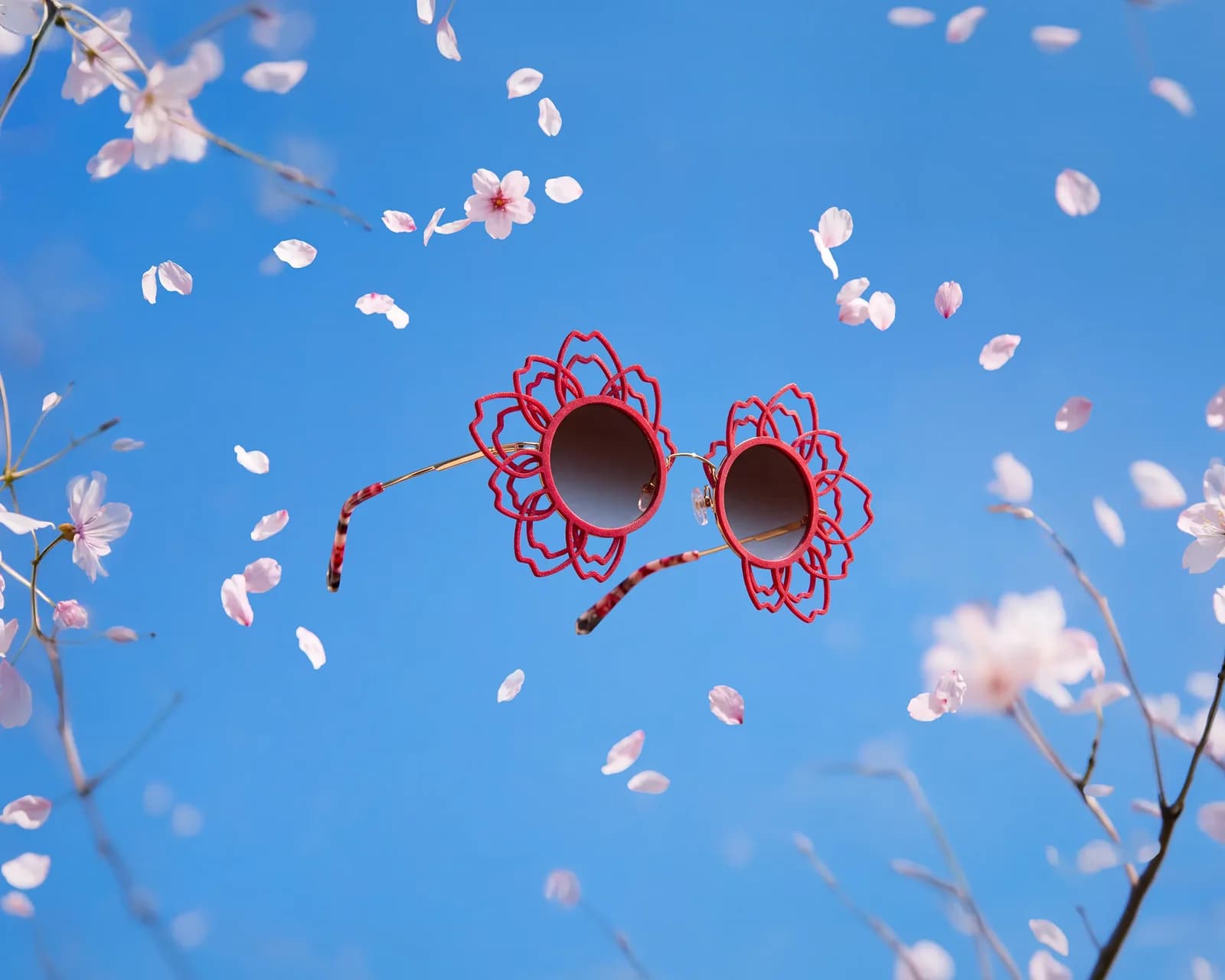J.F. REY SAKURA sunglasses with pink-red openwork petal rims, shown among cherry blossoms against blue