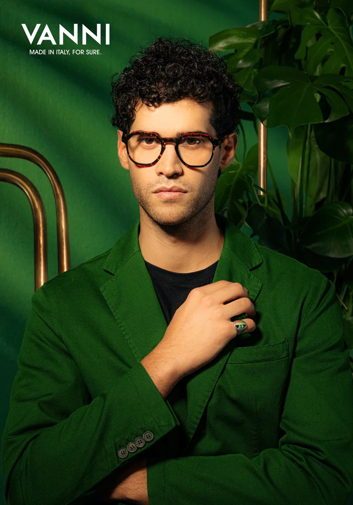 Man in a green jacket wearing dark patterned VANNI optical frames with red and amber details against green foliage