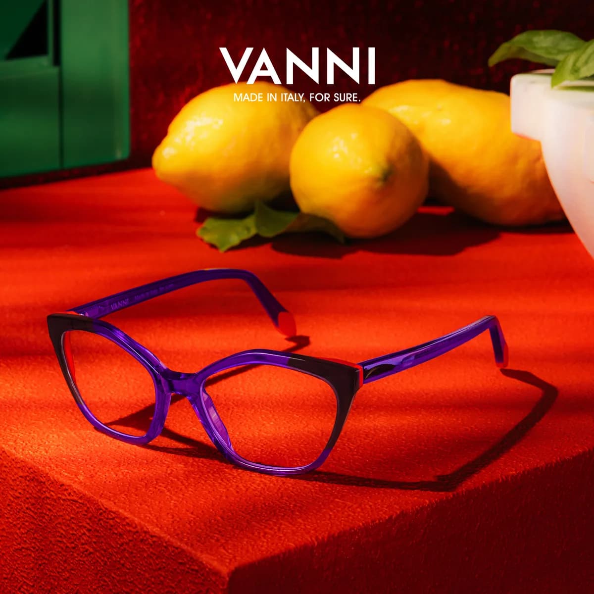 Angular violet VANNI optical frame with dark upper corners on a red surface beside yellow lemons