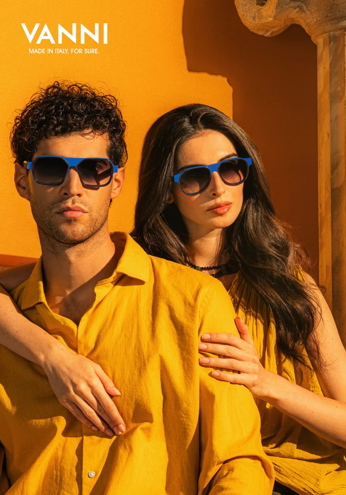 Two models wearing blue VANNI sunglasses and yellow clothing against a golden wall and stone column
