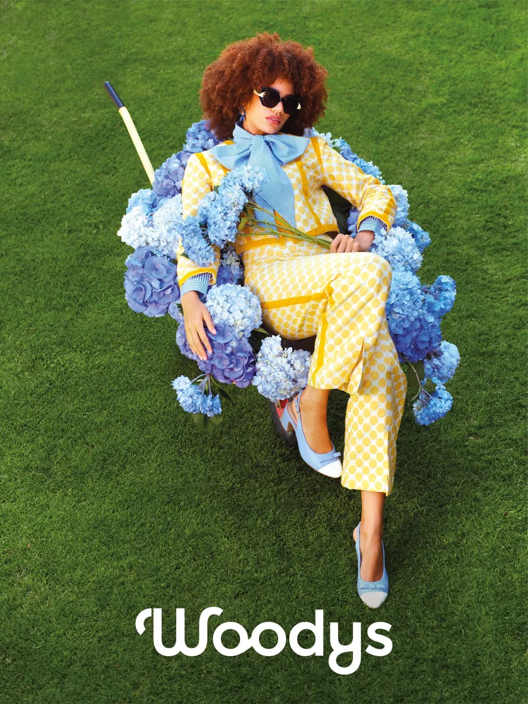 Model wearing Woodys Lea sunglasses in a wheelbarrow filled with blue flowers